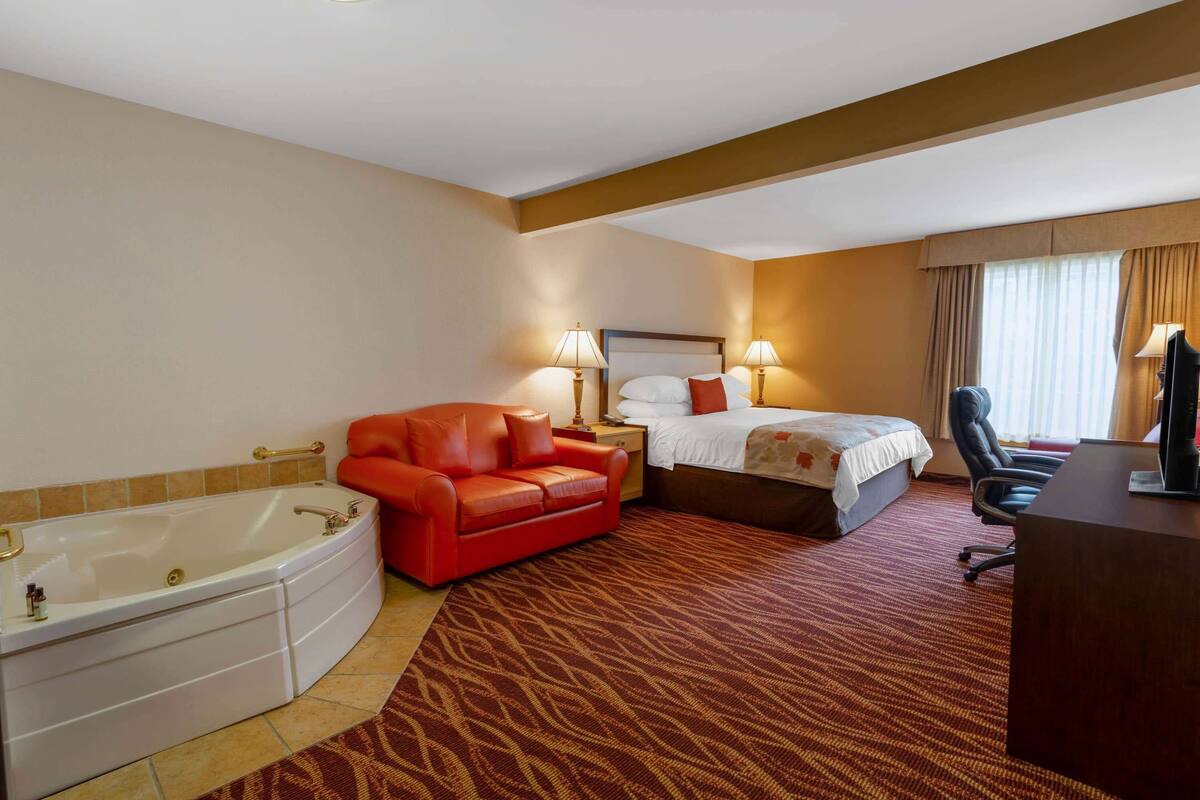 Ein Hotelzimmer mit einer großen Badewanne, einem roten Sofa, einem Bett mit weißen Leinen und einem Schreibtisch mit einem Computer.