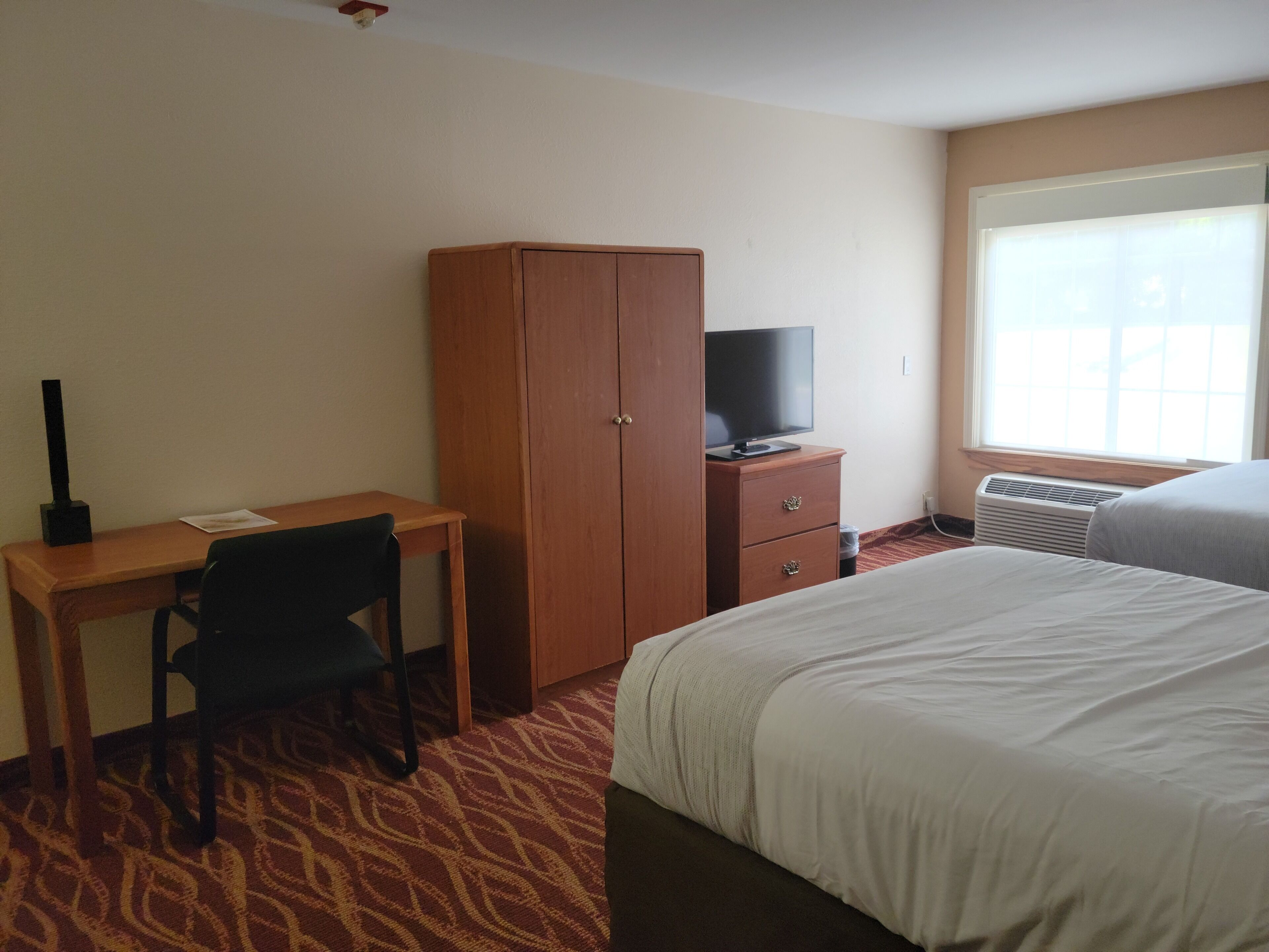 A hotel room with a bed, a desk with a chair, a wardrobe, a television on a dresser, and a window with blinds.