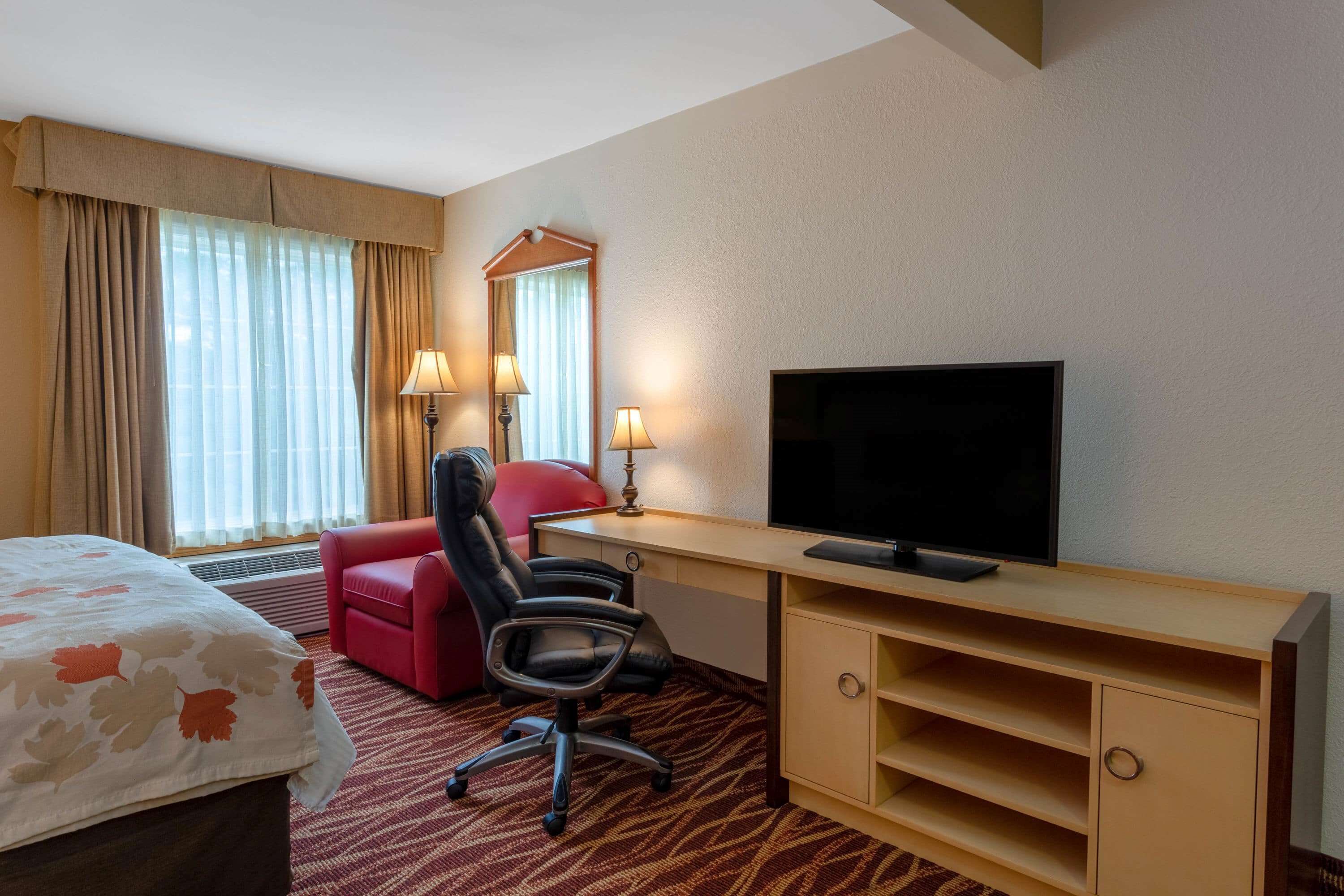 A hotel room with a bed, a desk with a television, a red chair, and a large window with curtains.