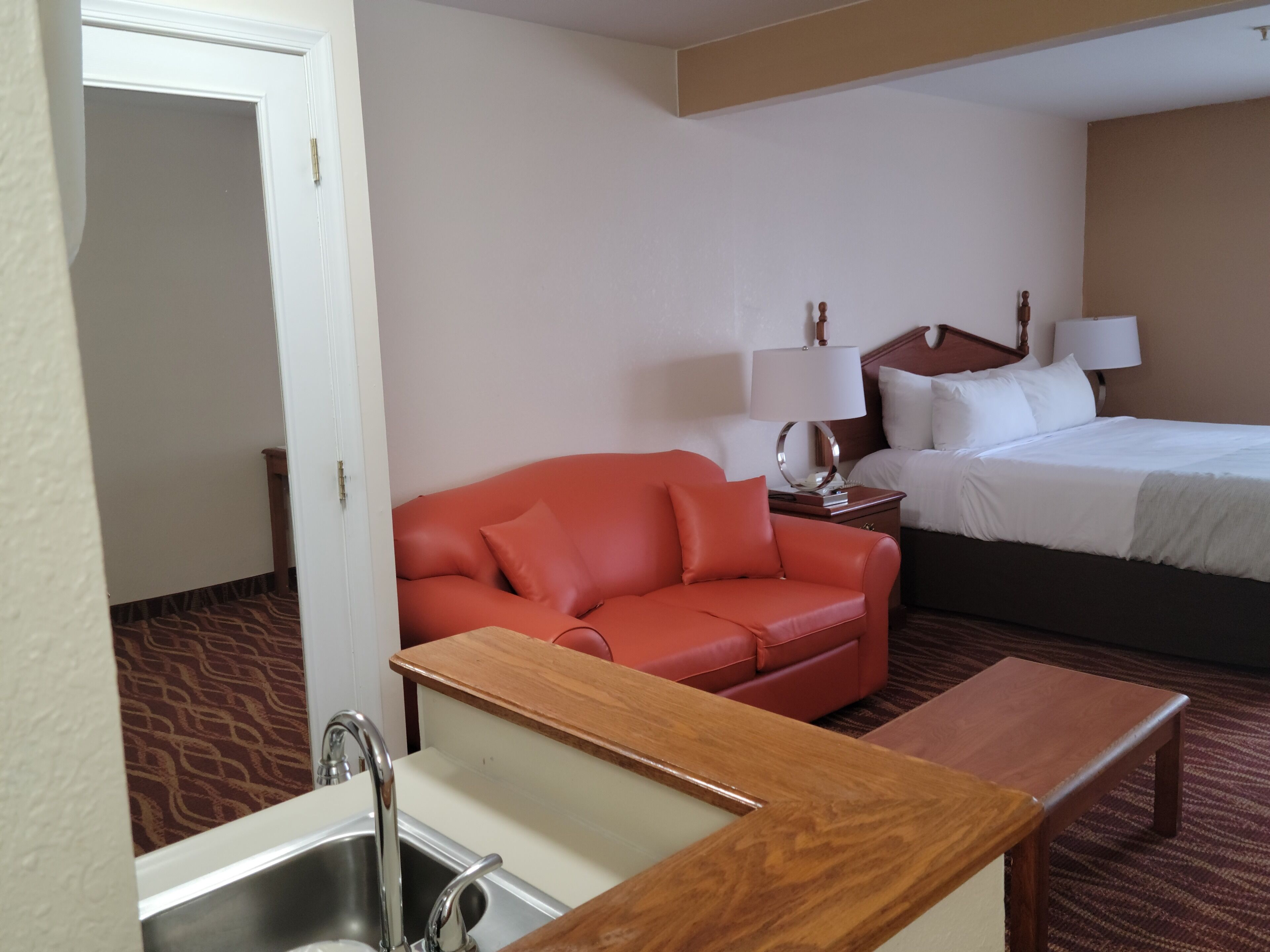 Una habitación de hotel con un sofá de cuero rojo, una cama con ropa de cama blanca, una mesita de noche de madera con lámparas y una pequeña mesa de madera.