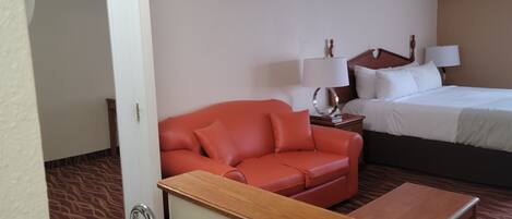 Una habitación de hotel con un sofá de cuero rojo, una cama con ropa de cama blanca, una mesita de noche de madera con lámparas y una pequeña mesa de madera.