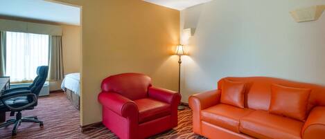 A hotel room with a red armchair, an orange sofa, a desk with a chair, a lamp, and a bed.