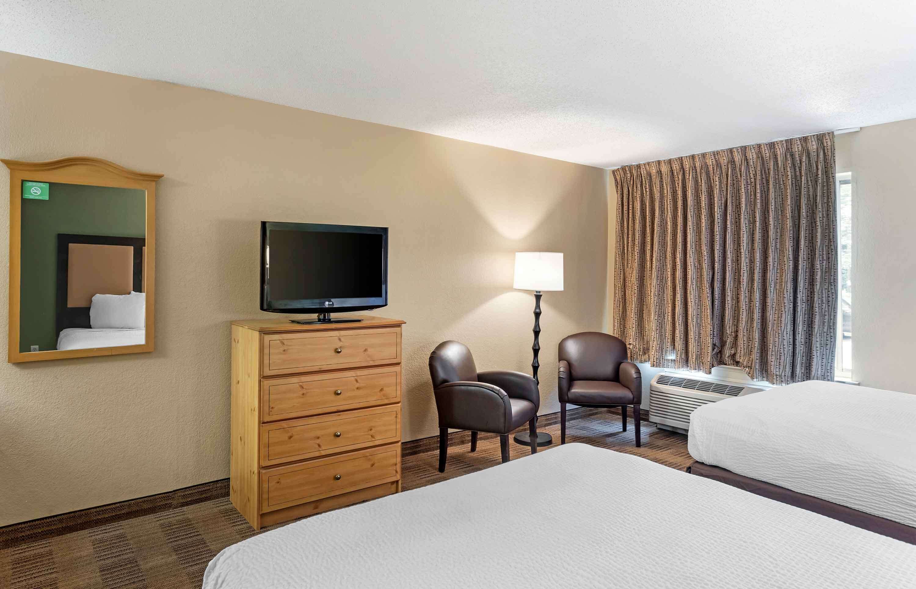 Une chambre d’hôtel avec deux lits, un téléviseur posé sur une commode en bois, une lampe, deux chaises et une fenêtre avec des rideaux.