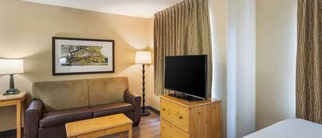 Una habitación de hotel con sofá, una mesa de madera, televisor sobre un mueble de madera, lámpara y cama.