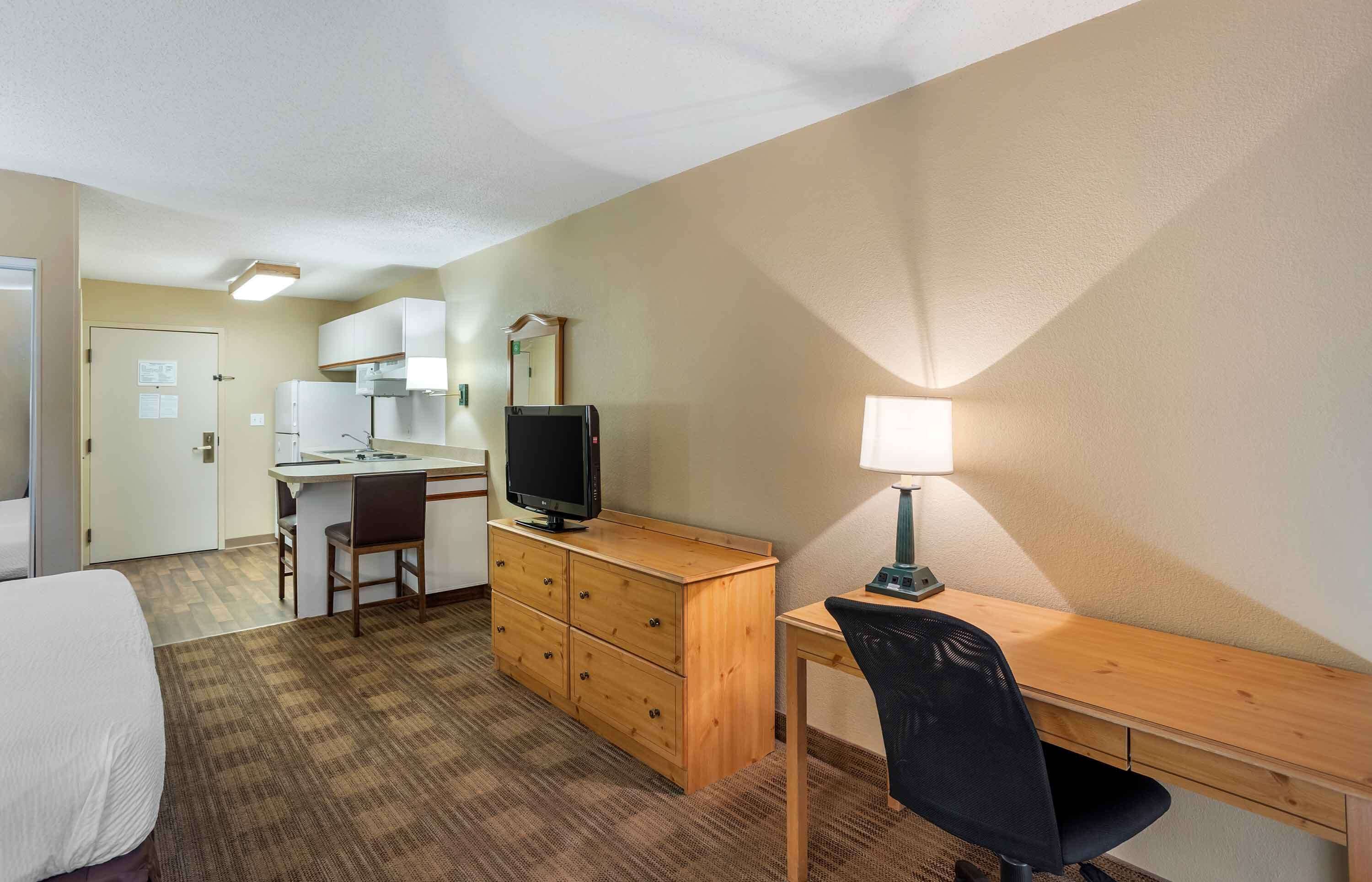 A hotel room with a bed, a desk with a chair, a television, a desk lamp, a dresser, and a kitchenette.