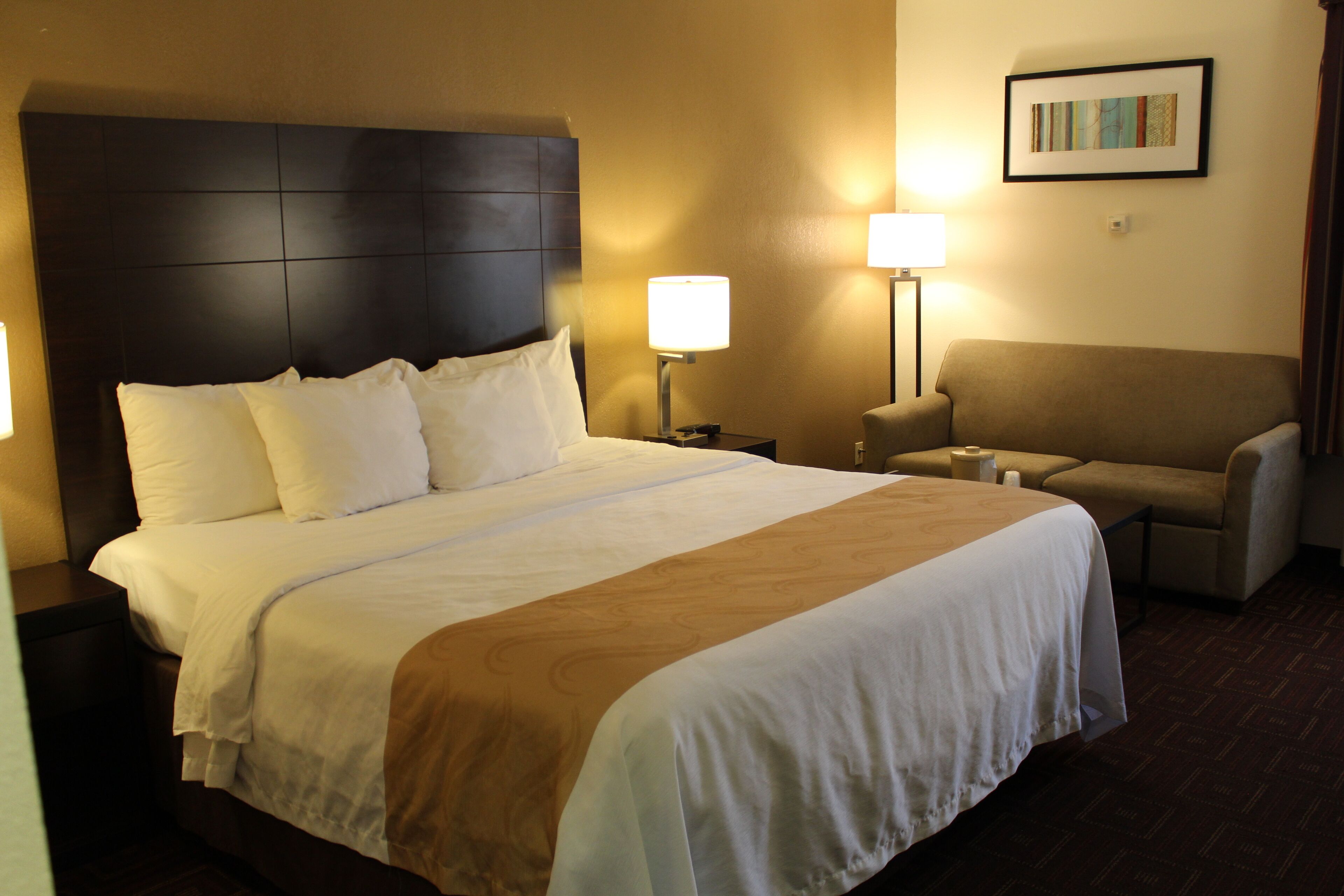 Quarto de hotel com uma cama grande, cabeceira, mesa de cabeceira, abajur, uma cadeira e um quadro na parede.