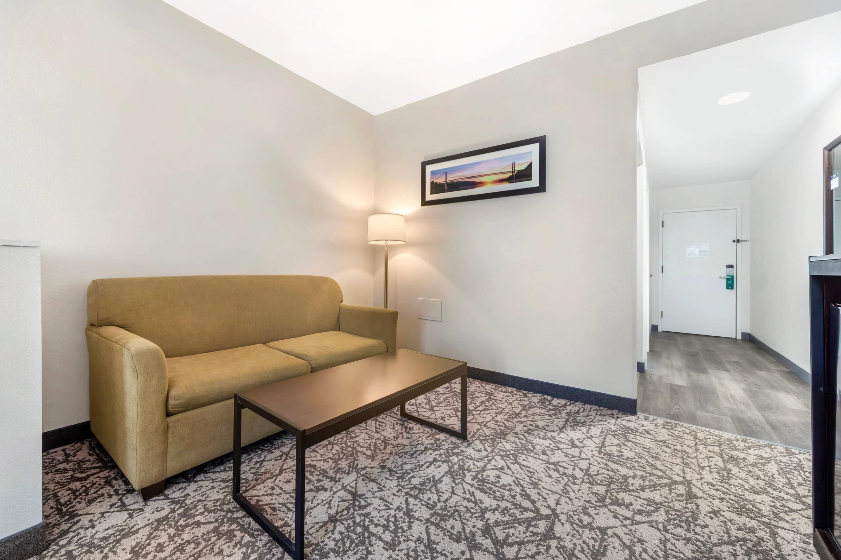 A hotel room with a beige sofa, a small table, a patterned rug, a framed picture on the wall, and a hallway leading to a door.