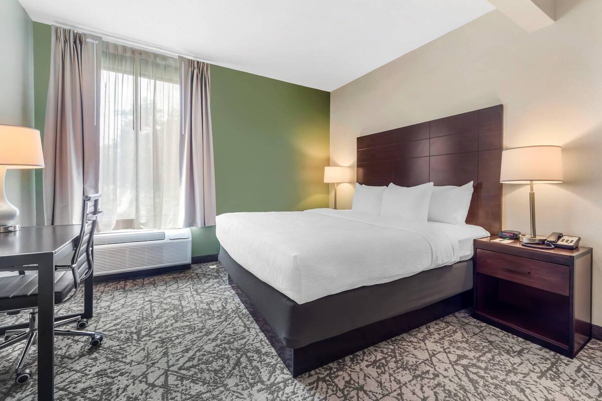 A hotel room with a large bed, bedside lamps, a desk with a chair, and a window with curtains.
