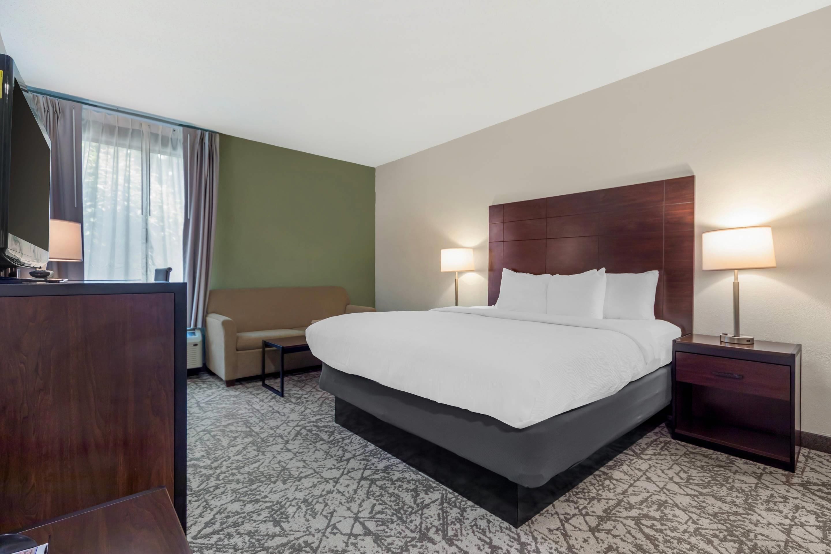 A hotel room with a large bed, two bedside lamps, a wooden nightstand, a desk with a lamp, a sofa, and a view of the outdoors through curtains.