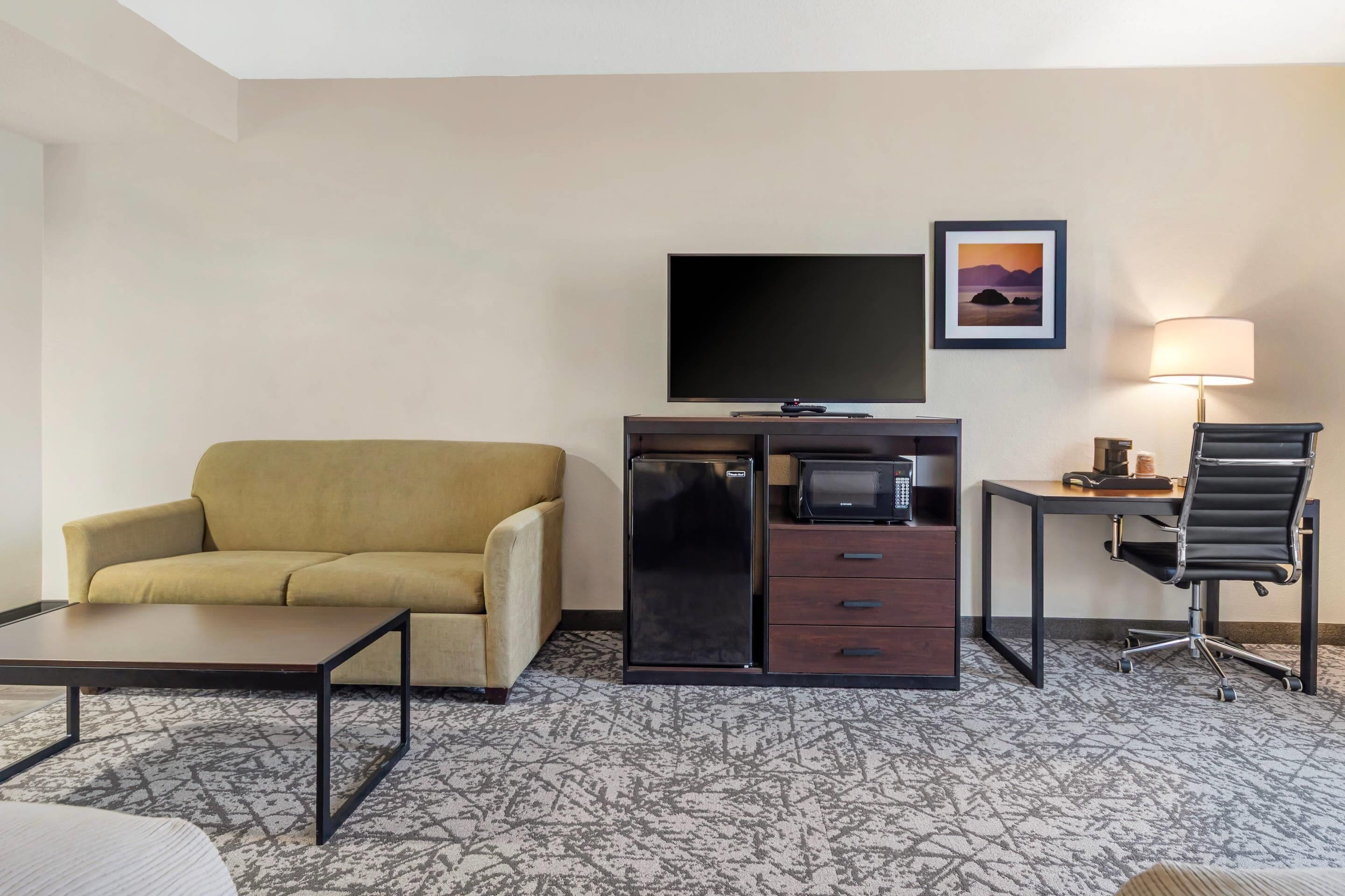 Habitación de hotel con sofá, televisor de pantalla plana, refrigerador, microondas, escritorio con lámpara y una silla.