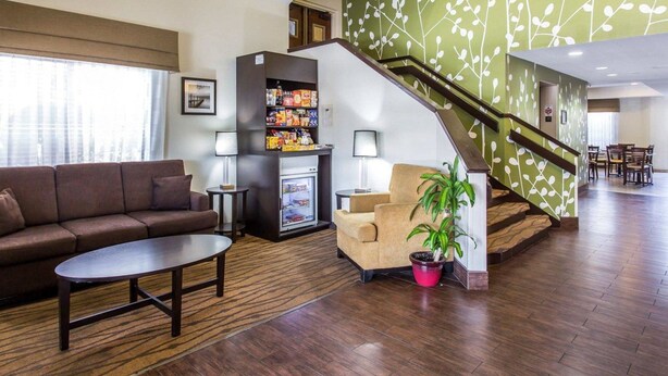 Um lobby de hotel com um sofá marrom, uma mesinha, um frigorífico cheio de lanches, uma escada com uma parede verde e uma área de refeições ao fundo.
