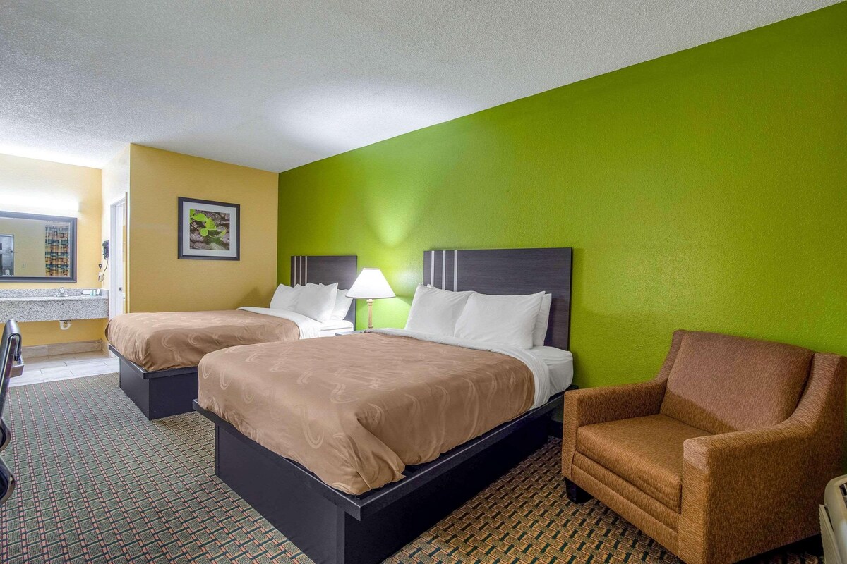 A hotel room with two beds, a green wall, a brown armchair, and a patterned carpet.