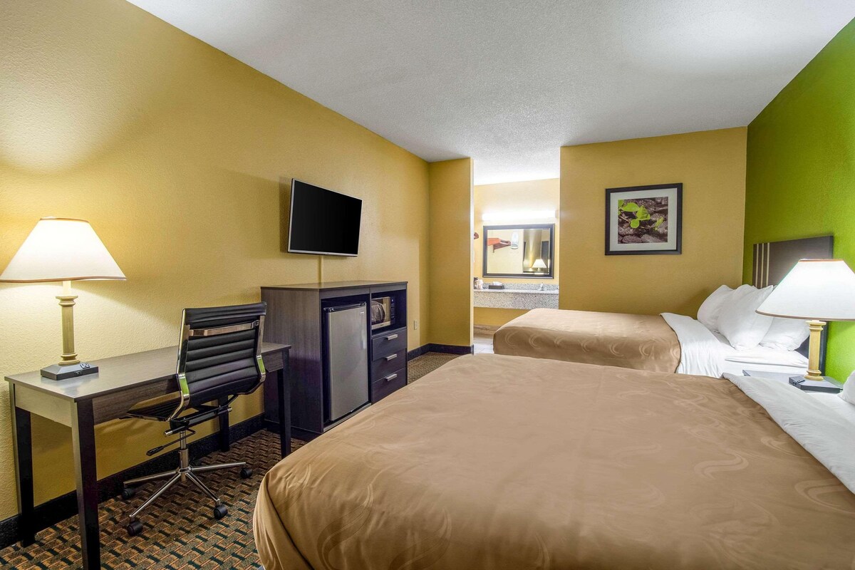 A hotel room with a desk, chair, television, two beds, and a bathroom mirror.