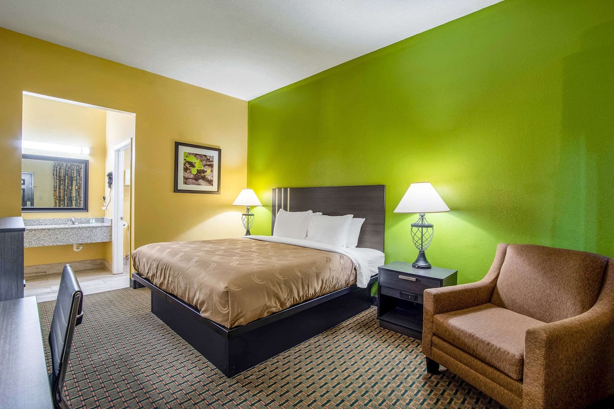 A hotel room with a bed, two bedside lamps, a chair, a desk, and a bathroom visible through an open door.