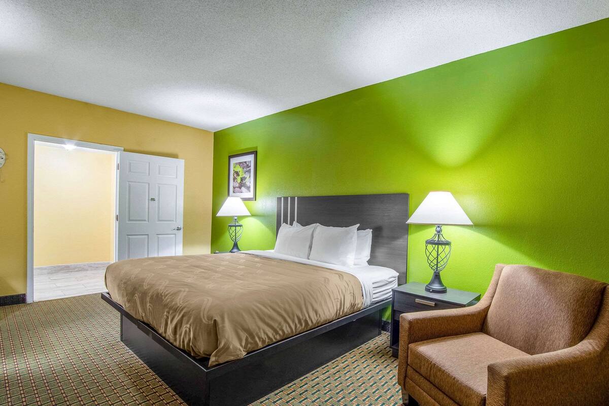 A hotel room with a bed, a chair, a nightstand with lamps, and a door.