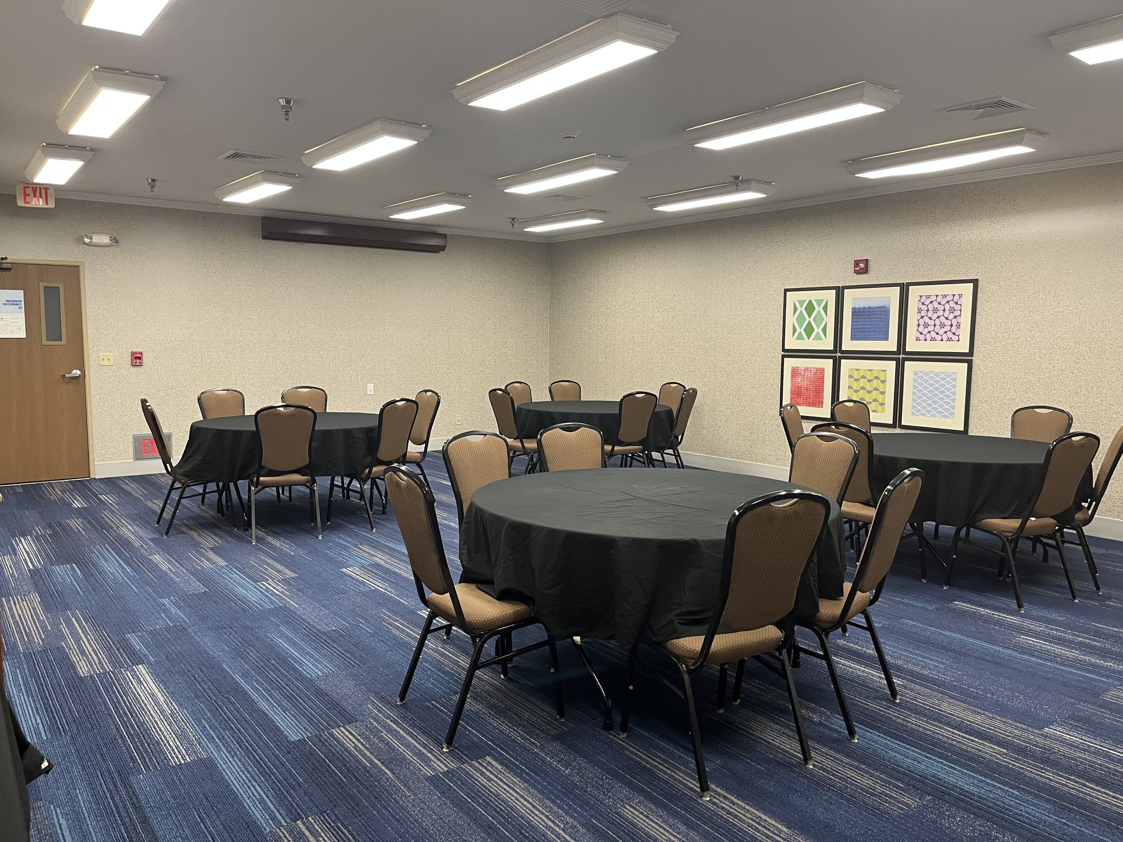 Une salle de conférence avec des tables et des chaises rondes, un sol recouvert de moquette et des luminaires au plafond.