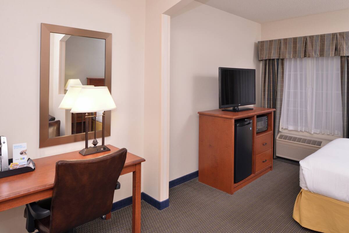 A hotel room with a wooden desk, a chair, a television on a cabinet, a bed, a window with curtains, and a mirror on the wall.