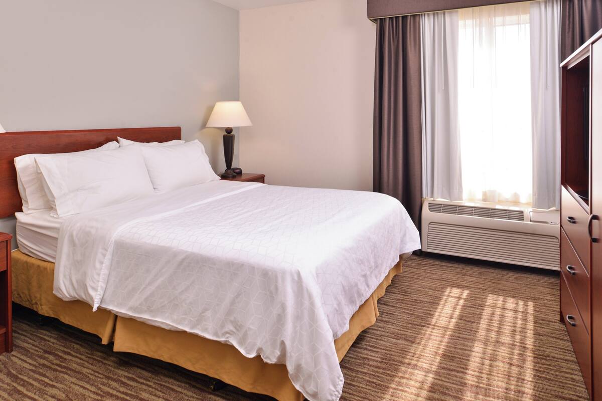 A hotel room with a bed, a nightstand, a lamp, an air conditioning unit, and a window with curtains.