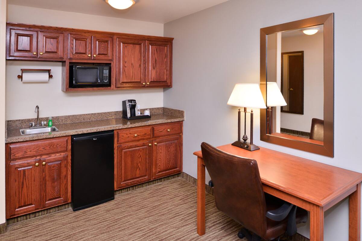 A compact kitchen with wooden cabinets, a microwave, a coffee maker, and a desk with a chair.
