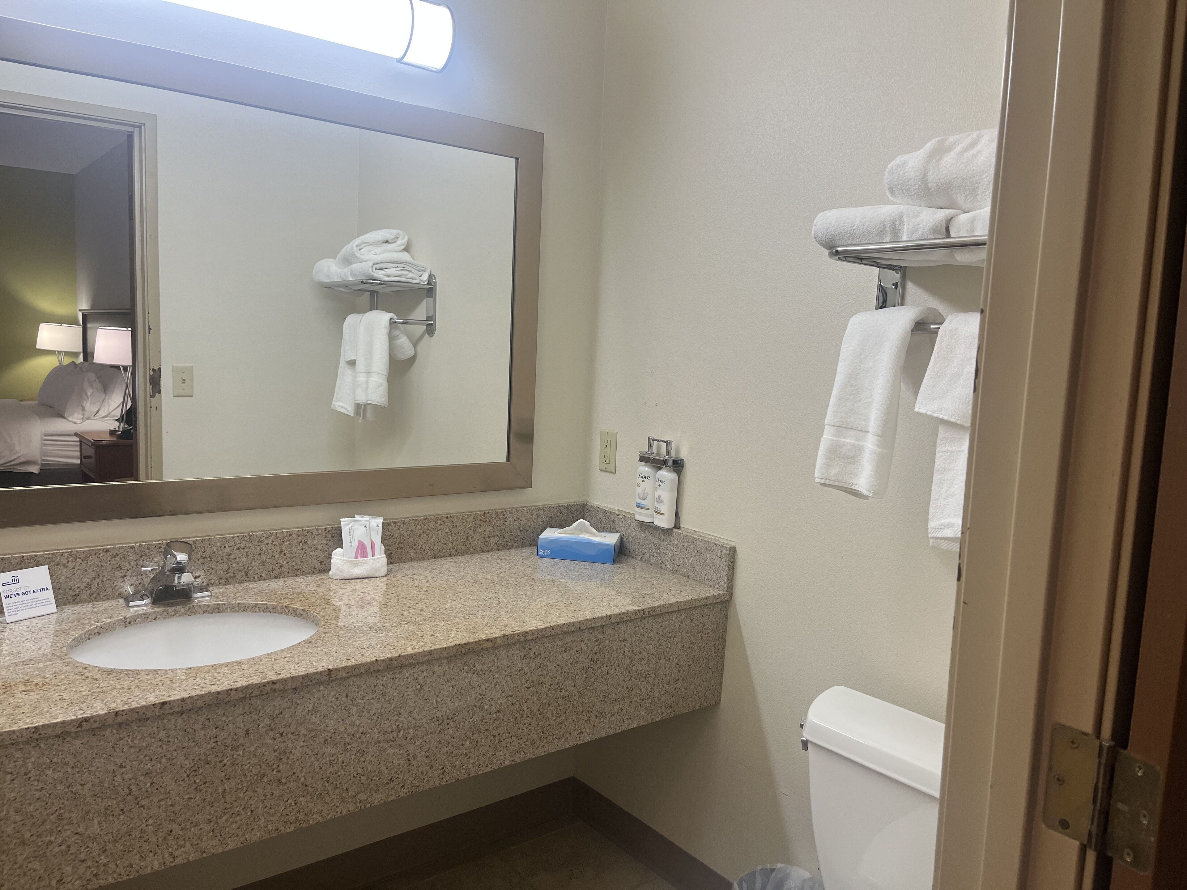 A hotel bathroom with a granite countertop, a sink, a mirror, a toilet, and a towel rack.