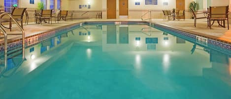 An indoor swimming pool with clear water, surrounded by lounge chairs and tables.