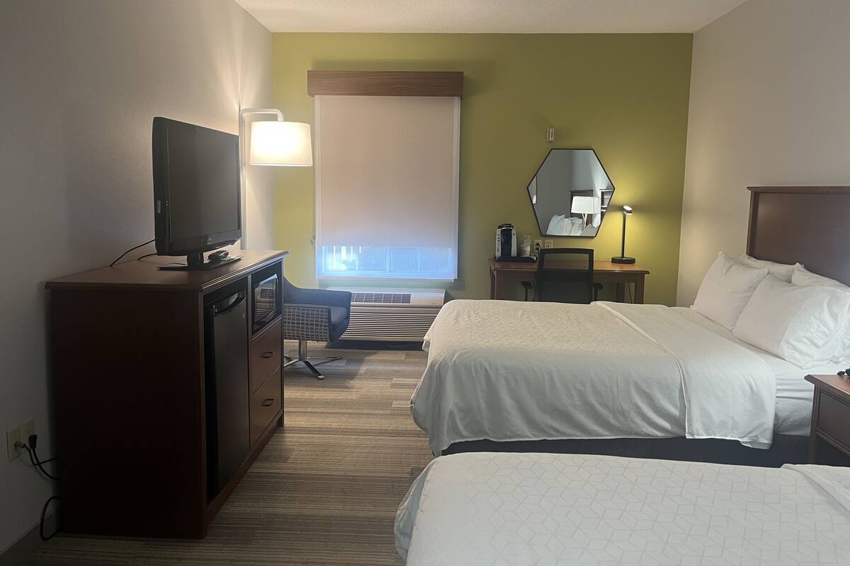 A hotel room with two beds, a TV, a desk with a lamp, and a large window with blinds.