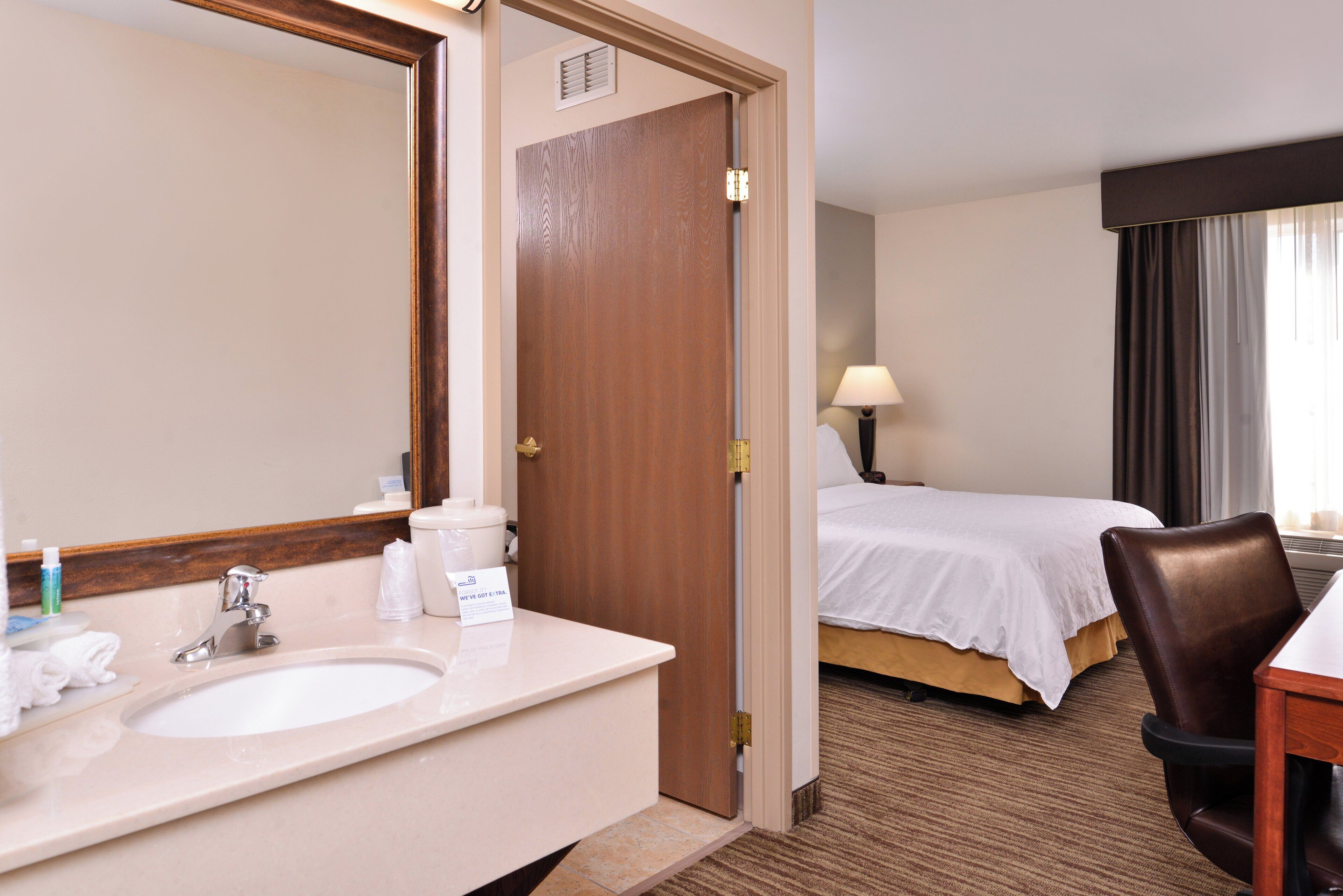 A hotel room with a bathroom featuring a sink, mirror, and toiletries. There is a bed with a white comforter and a brown chair.