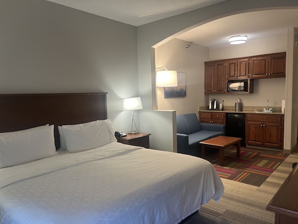 A hotel room with a large bed, a small desk, a chair, a kitchenette, and a nightstand with a lamp.