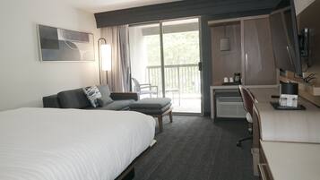 A hotel room with a large bed, a sofa, a desk, and a balcony with a view of trees.
