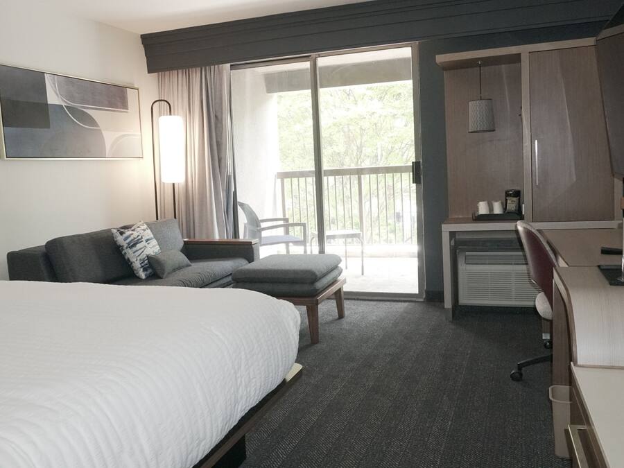 A hotel room with a large bed, a sofa, a desk, and a balcony with a view of trees.