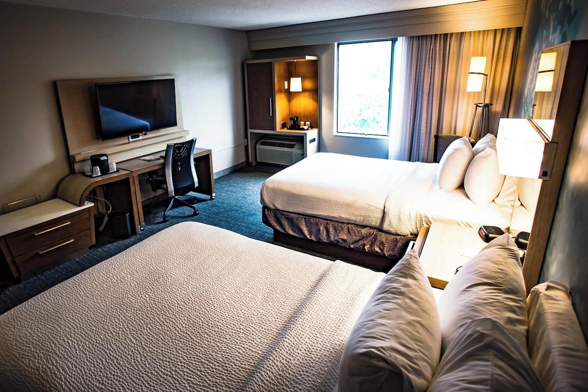 A hotel room with two beds, a TV, a desk with a chair, a desk lamp, and a window with curtains.