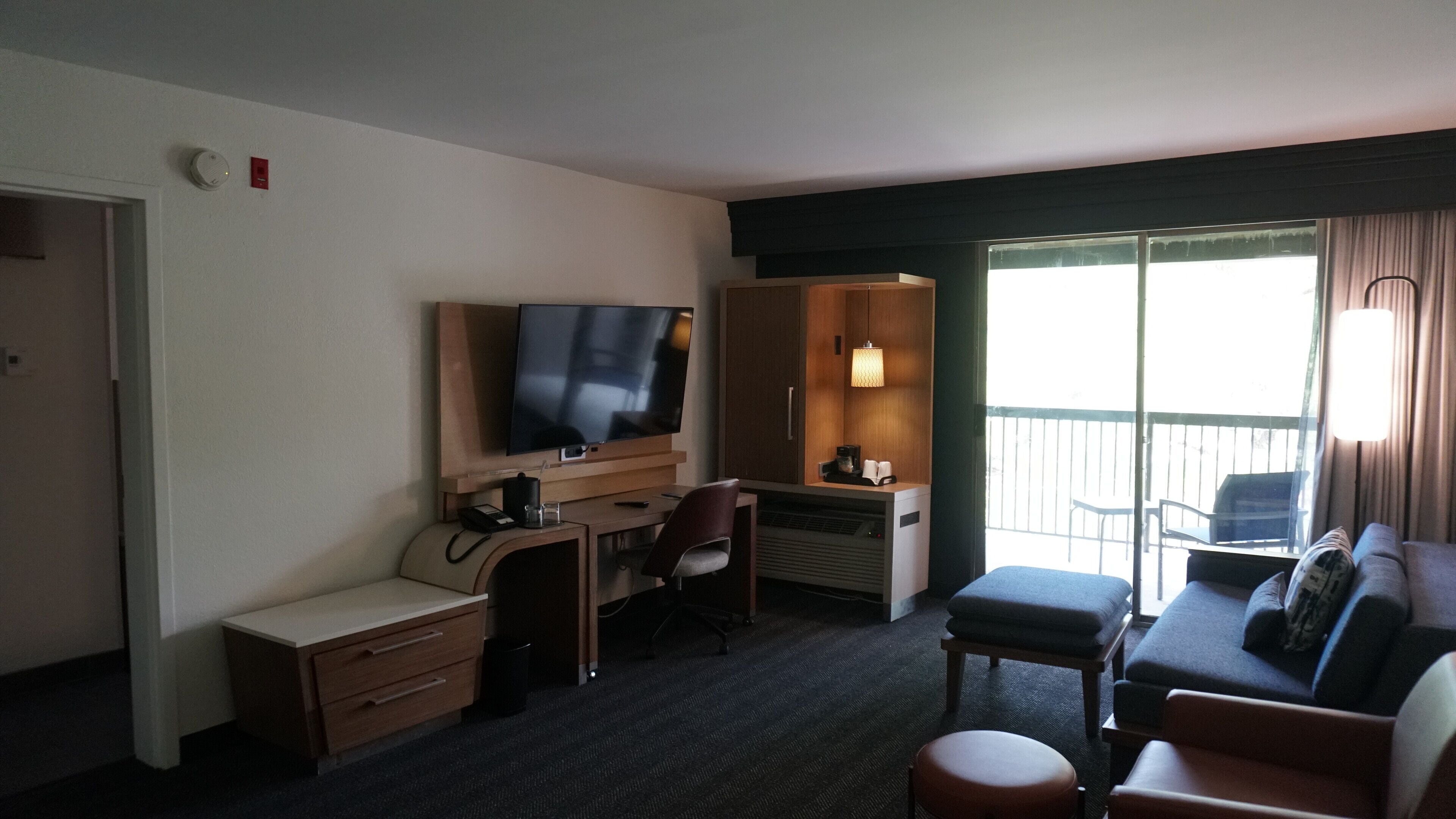 A hotel room with a flat-screen TV, a desk with a chair, a sofa, and a balcony with a view.