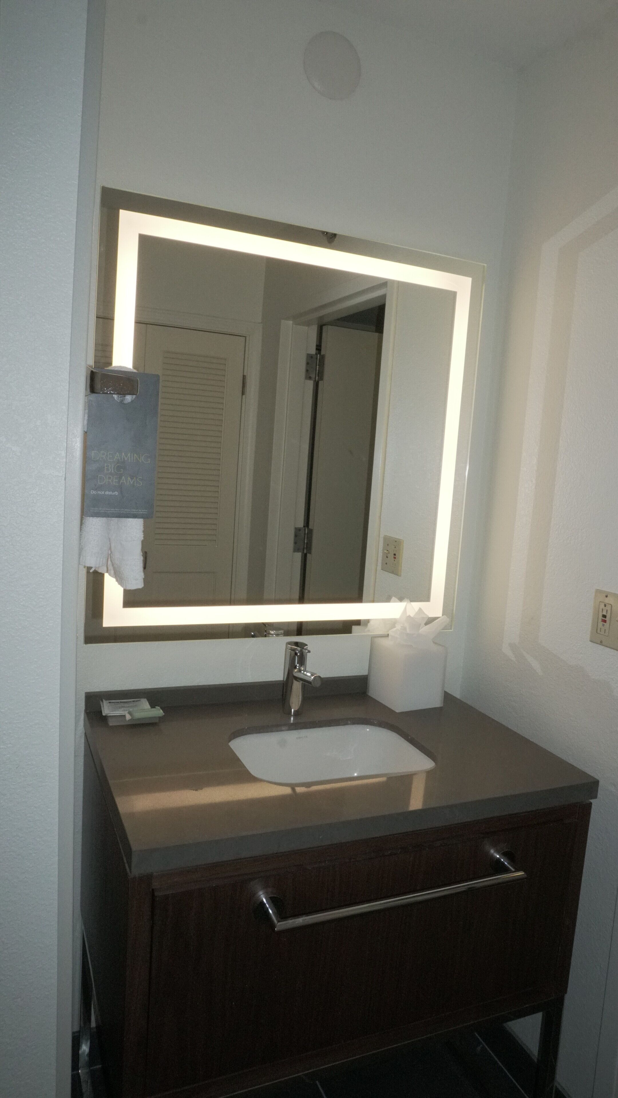 A bathroom with a large mirror, a sink, a towel dispenser, and a cabinet.