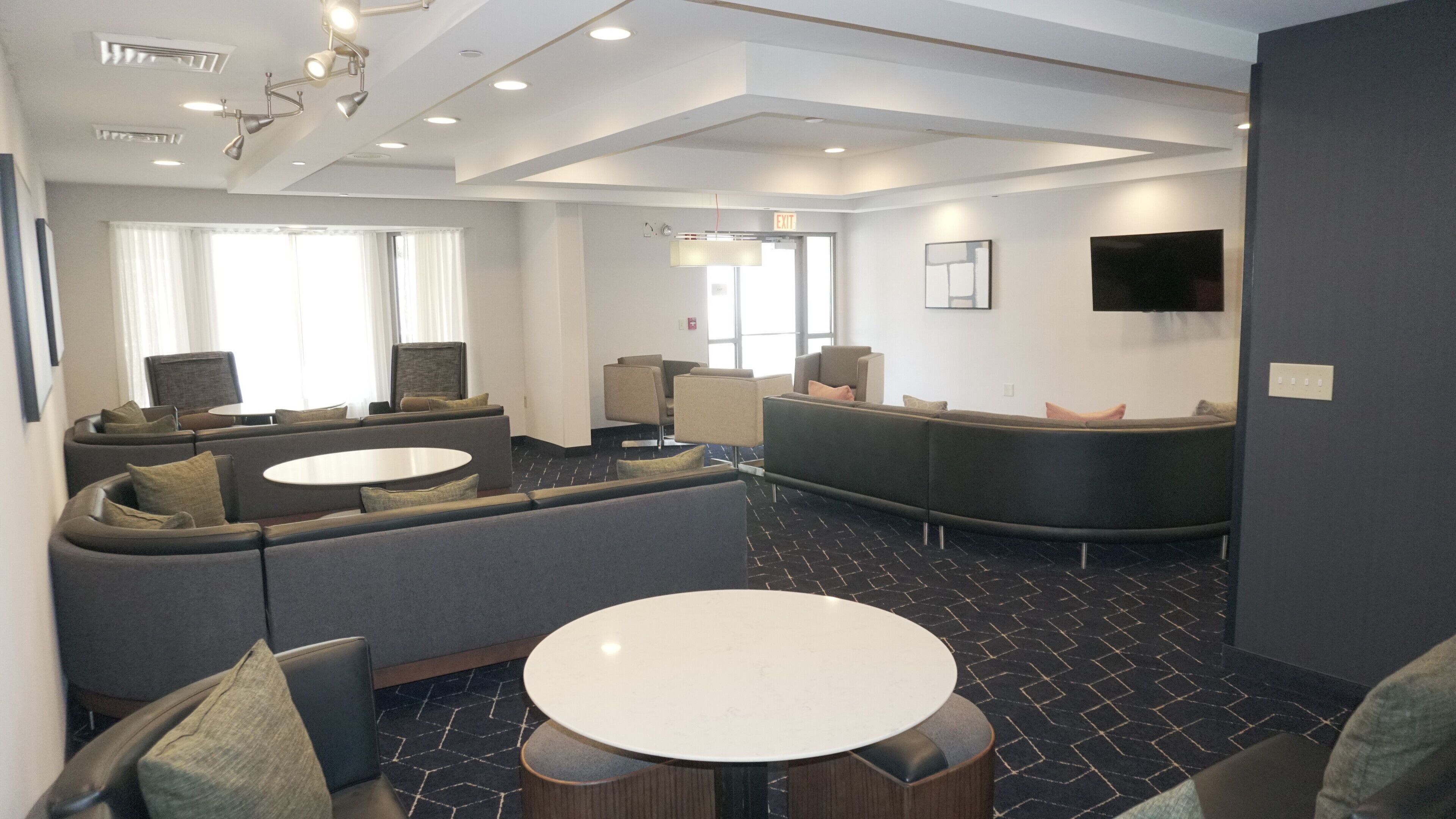 A modern hotel lobby with a curved reception desk, seating area with sofas and chairs, a round table, and a flat-screen TV mounted on the wall.