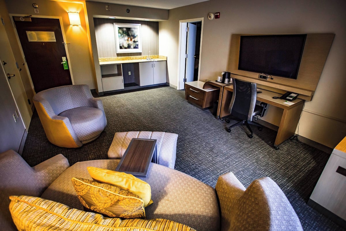 A hotel room with a sofa, armchair, desk, TV, and a small kitchenette area.