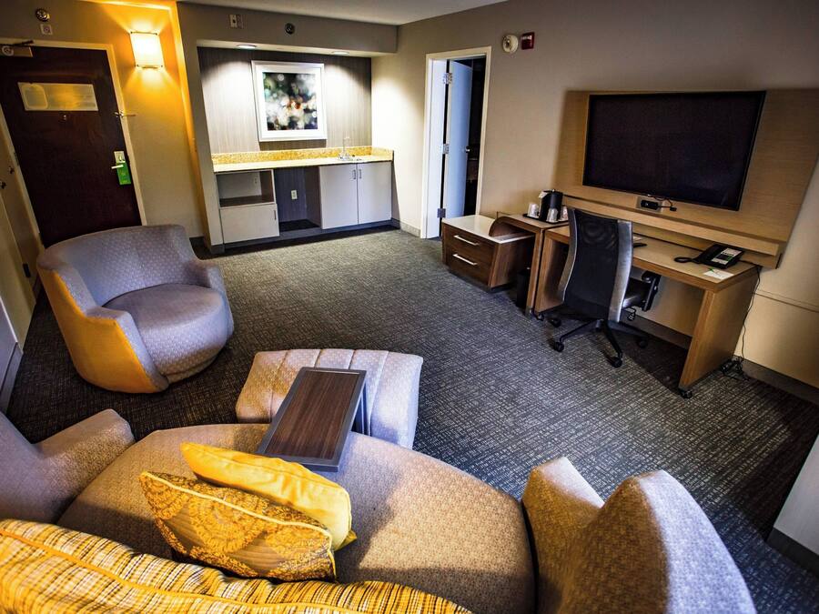 A hotel room with a sofa, armchair, desk, TV, and a small kitchenette area.