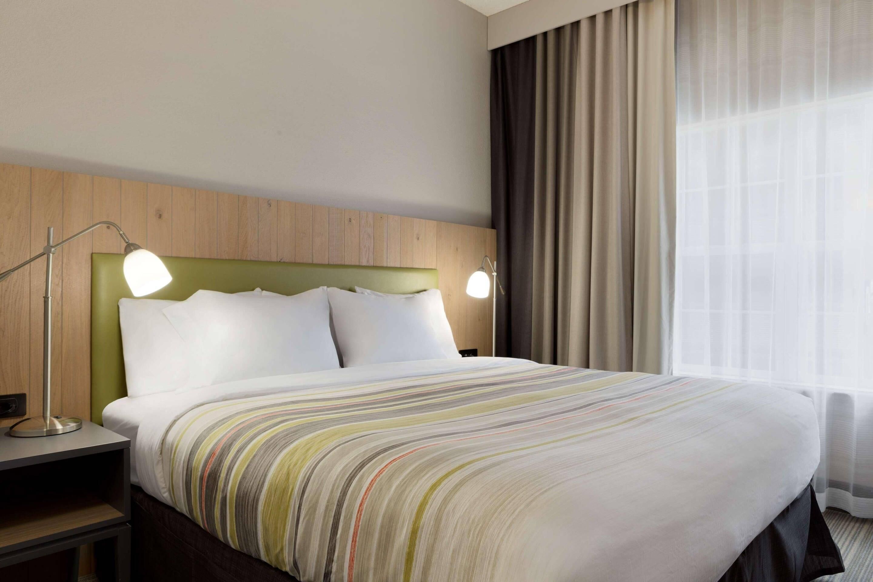 A hotel room with a neatly made bed, two bedside lamps, a wooden headboard, and a large window with curtains.