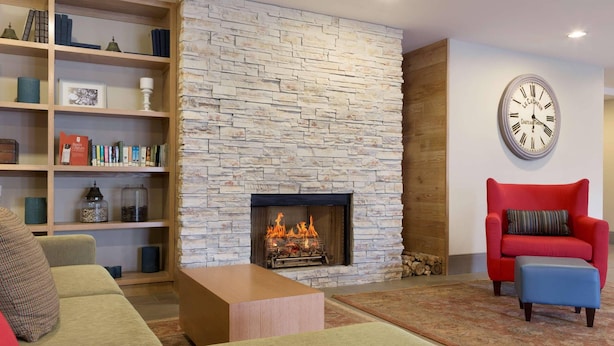 A living room with a stone fireplace, a bookshelf, a red armchair, and a blue ottoman. - Country Inn & Suites by Radisson, Jackson, TN (Jackson)