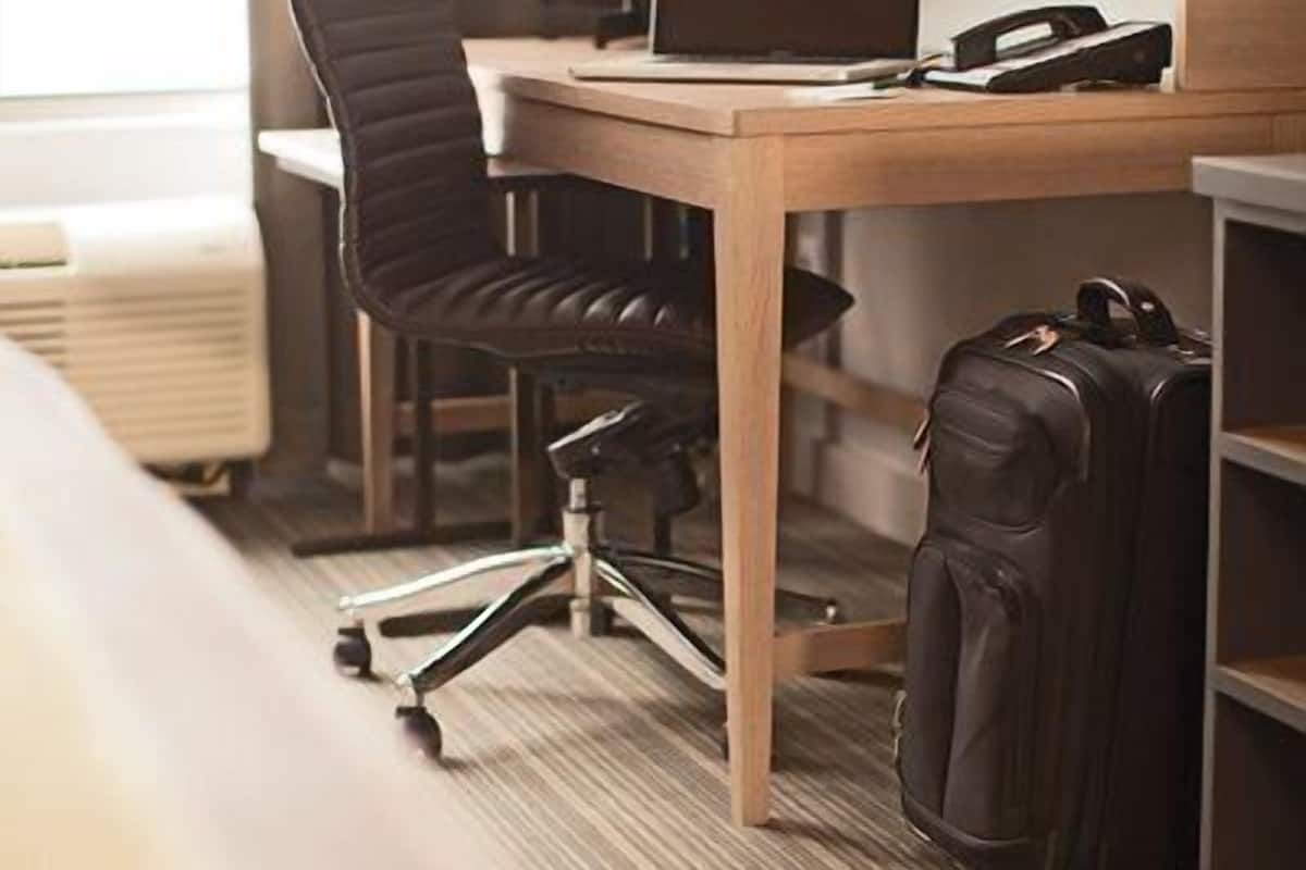 A hotel room with a wooden desk, a black office chair, a laptop, a lamp, and a suitcase.