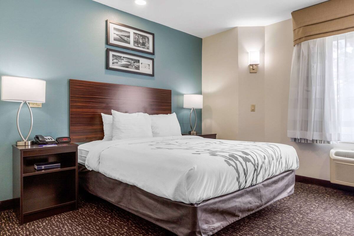 Una habitación de hotel con una cama grande, dos lámparas de noche, una mesita de noche con un teléfono y un libro, y cuadros en la pared.
