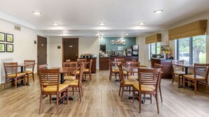 Un espace repas avec des tables et des chaises en bois, une cuisine à l’arrière-plan et de grandes fenêtres.