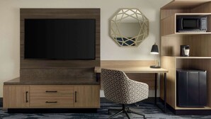 A modern hotel room with a flat-screen TV mounted on a wooden cabinet, a geometric mirror on the wall, a patterned office chair, a desk with a lamp, and built-in appliances including a microwave and refrigerator. - Fairfield Inn & Suites by Marriott Austin Southwest (Austin)