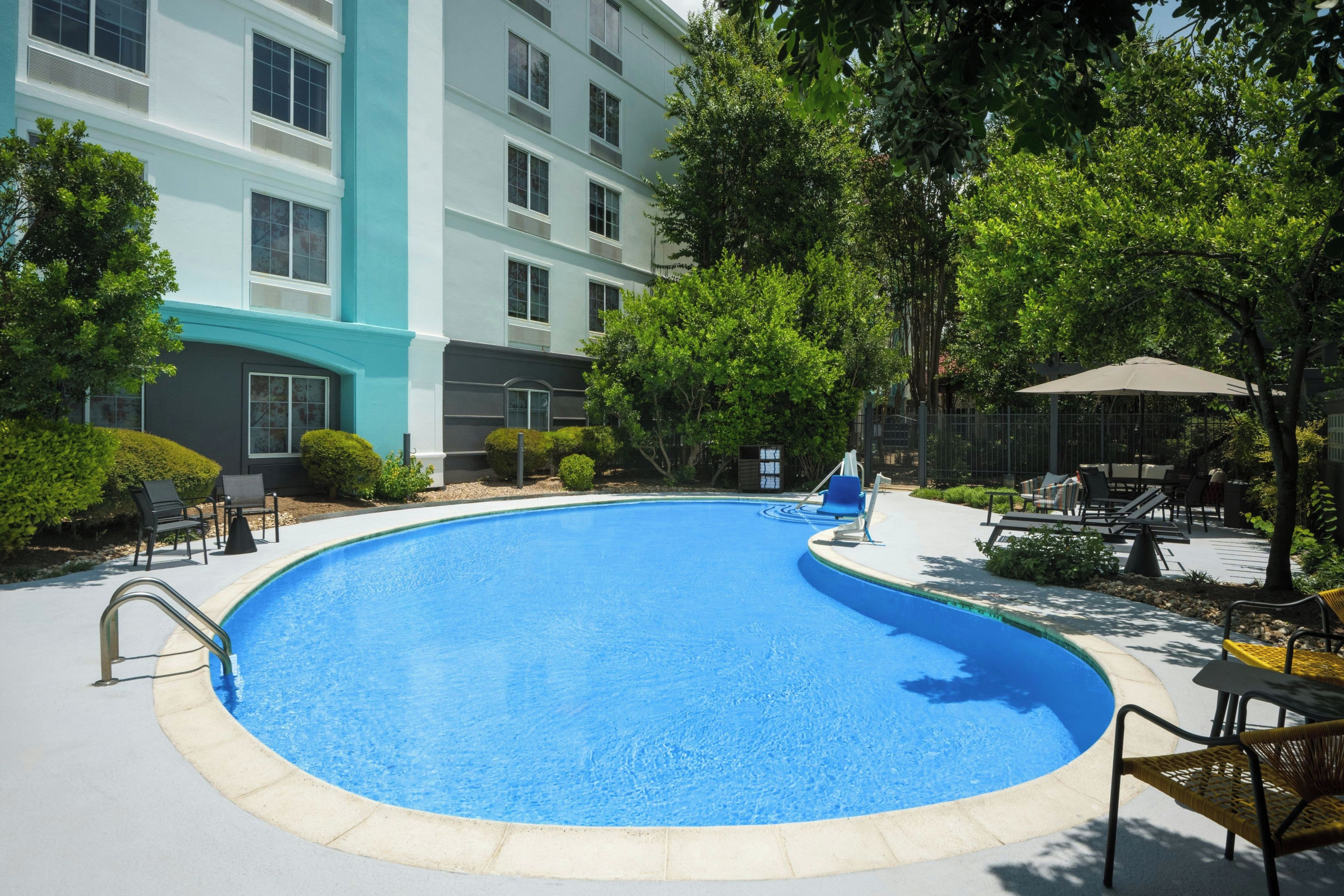 A swimming pool with lounge chairs and umbrellas, surrounded by greenery and a building with multiple windows.
