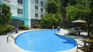 A swimming pool with lounge chairs and umbrellas, surrounded by greenery and a building with multiple windows.
