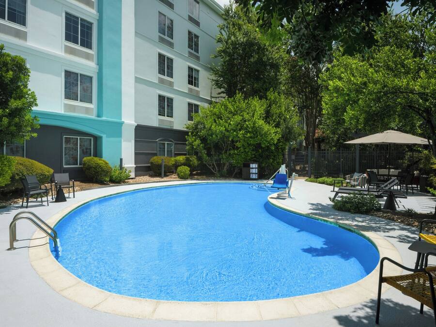 A swimming pool with lounge chairs and umbrellas, surrounded by greenery and a building with multiple windows.