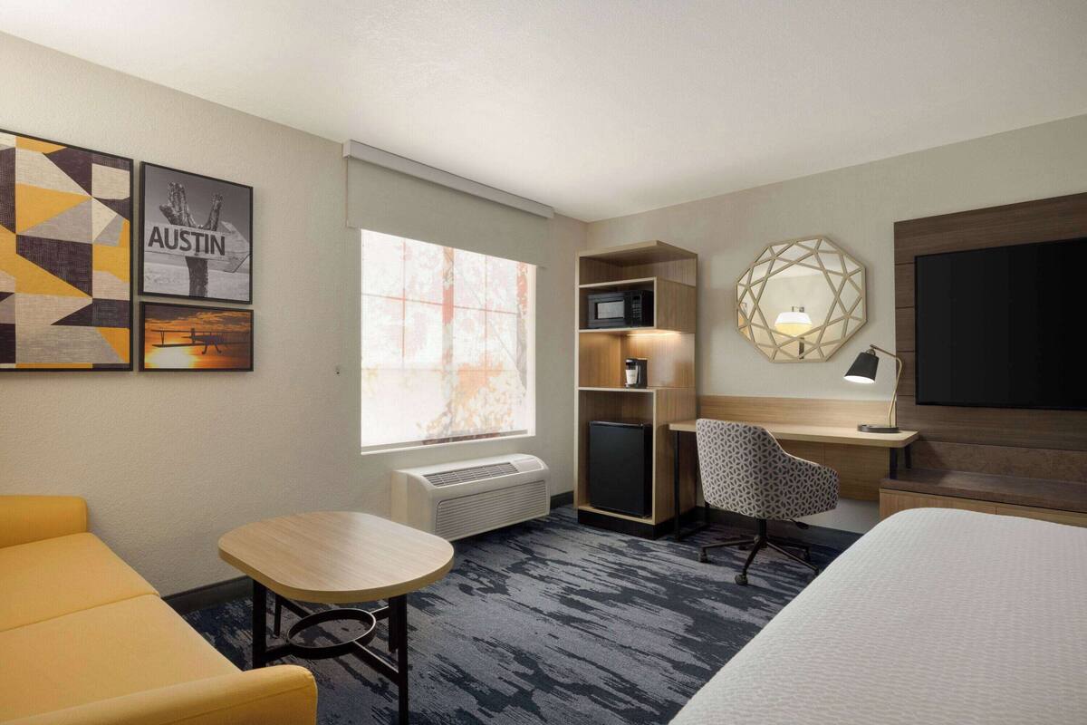 A hotel room with a bed, a desk with a computer, a chair, a microwave, a small fridge, and a TV.