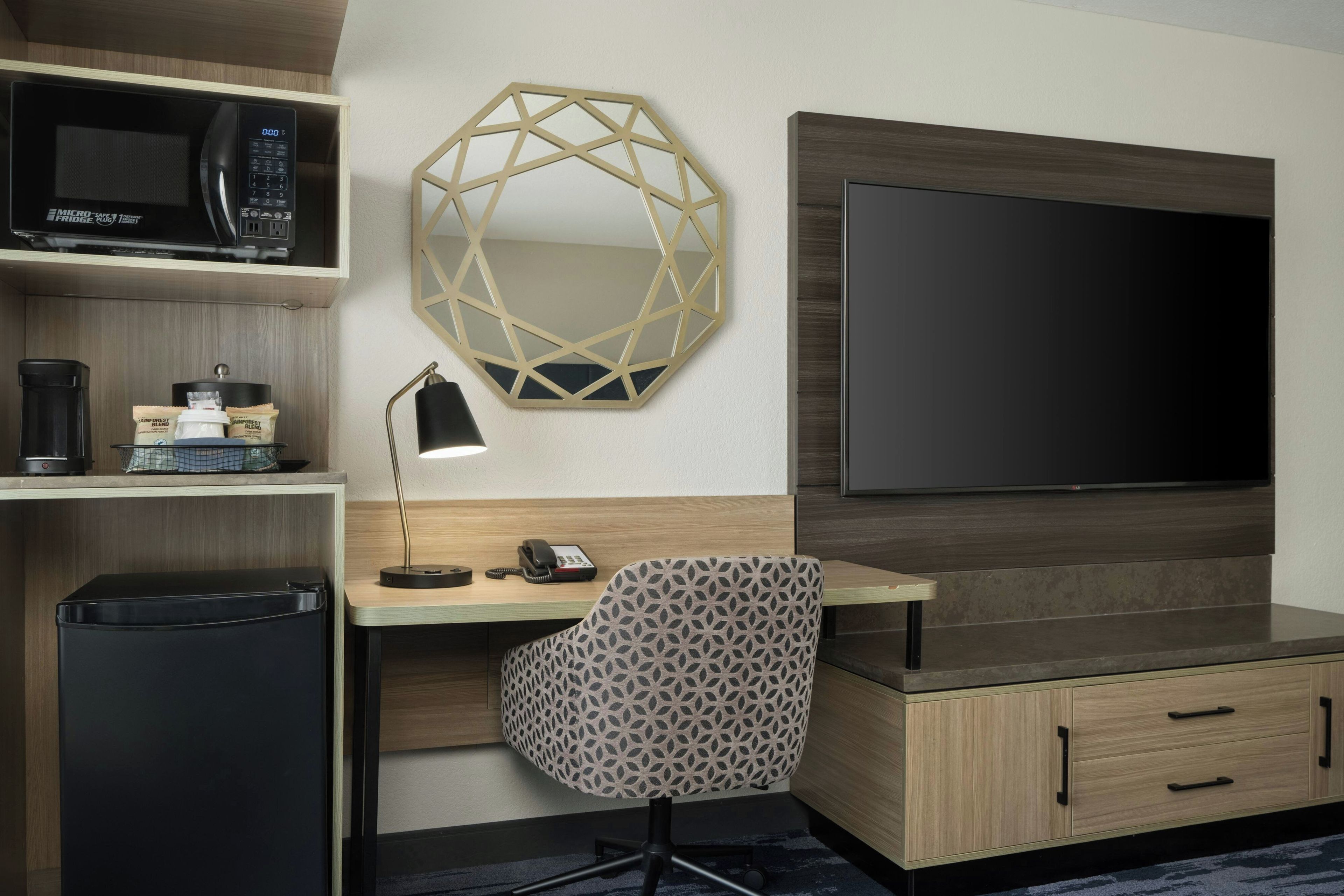 Quarto de hotel com cozinha compacta, micro-ondas, máquina de café, cadeira estampada, escrivaninha, telefone e televisão de tela plana.