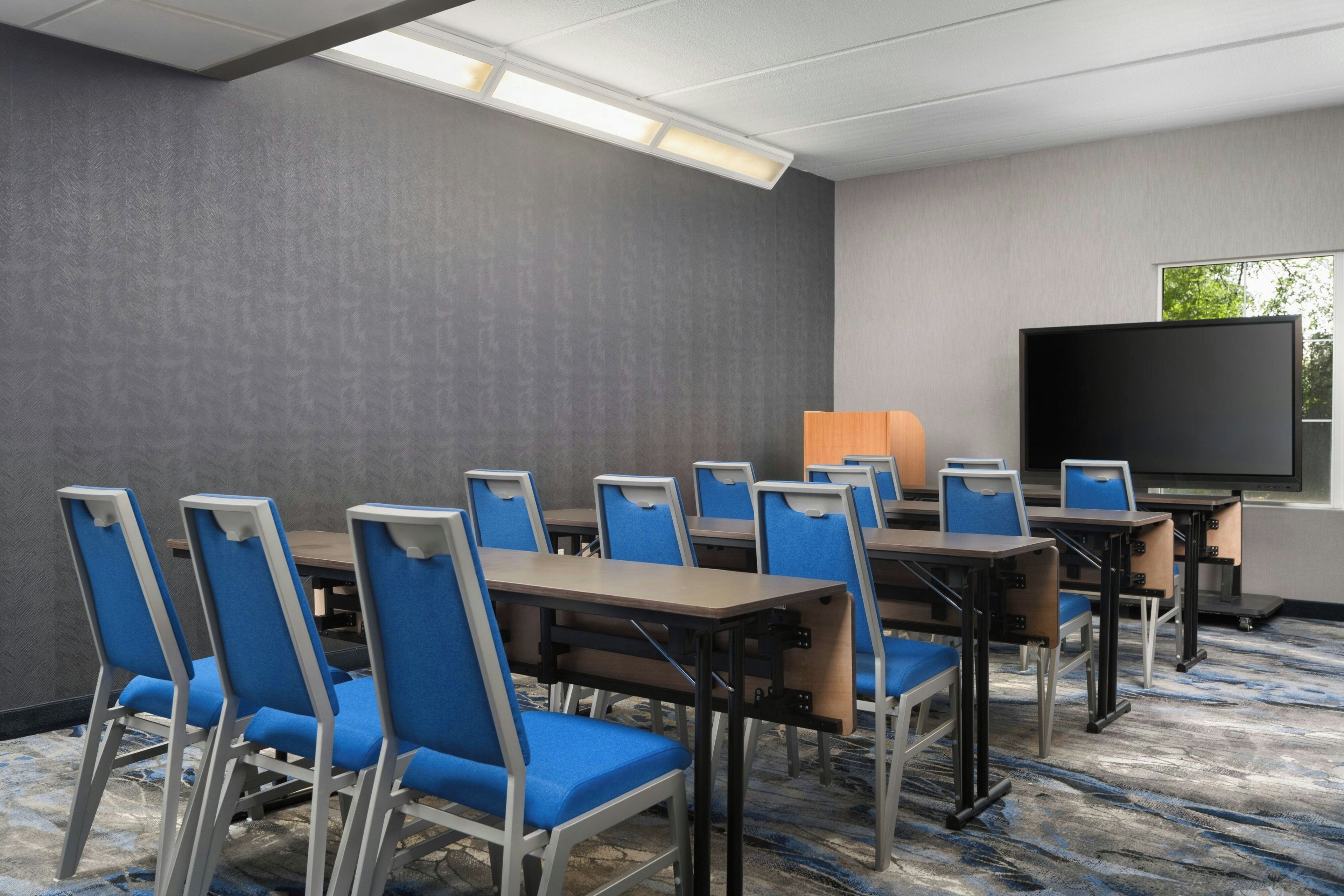 A conference room with multiple tables and chairs, a large flat-screen TV, and a window with a view of trees.