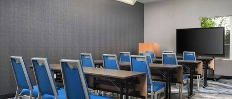 A conference room with multiple tables and chairs, a large flat-screen TV, and a window with a view of trees.