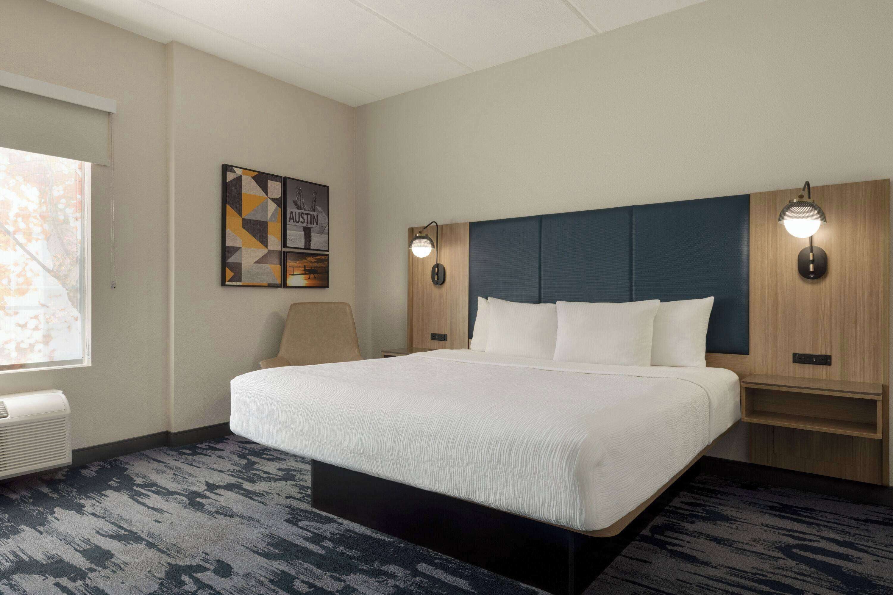 A hotel room with a large bed, a headboard, bedside tables, a wall-mounted lamp, a framed picture, and a window with blinds.